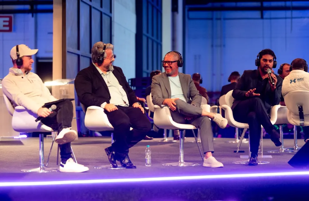 Quatro participantes com fones de ouvido sentados em palco durante painel de debate, um deles falando ao microfone